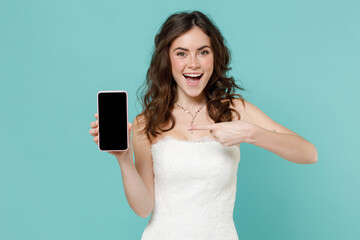 Excited bride young woman in beautiful white wedding dress pointing index finger on mobile cell phone with blank empty screen isolated on blue turquoise background. Ceremony celebration party concept.
