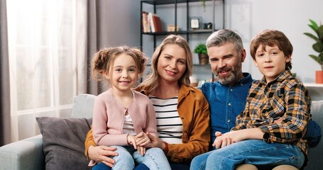 Portrait of cheerful positive Caucasian family parents with children gathered together at home sitting on sofa in room looking at camera and smiling in good mood, mom, dad with small daughter and son