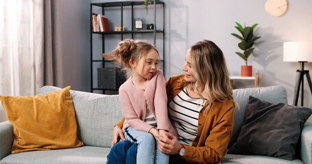 Portrait of Caucasian cheerful cute family mom and little daughter kid at home chatting having conversation while sitting on couch in modern living room, mom and child spending time at home together
