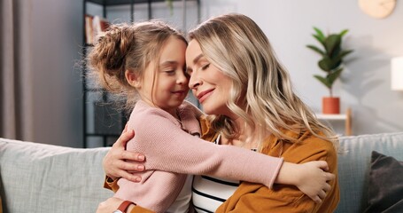 Close up of cute nice loving Caucasian little girl child hugging mother in living room at home. Caring mom embracing small daughter of sofa in apartment, love and care, family time concept