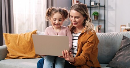 Portrait of happy nice Caucasian family daughter and mother spending time together at home using laptop online. Mom and cute small child girl tapping on computer surfing internet, leisure concept