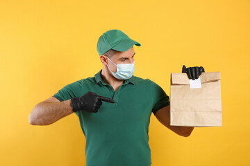Fototapeta na wymiar Courier in medical mask holding paper bag with takeaway food on yellow background. Delivery service during quarantine due to Covid-19 outbreak