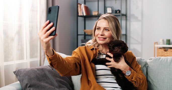 Close up portrait of positive beautiful Caucasian woman smiling posing to smartphone camera taking selfie photos with animal dog, joyful female with puppy pet taking pictures on cellphone at home