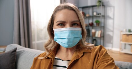 POV of joyful happy Caucasian beautiful female in medical mask speaking on video call waving hand spending time at home in quarantine. Close up of woman videochatting in living room. Covid-19 concept