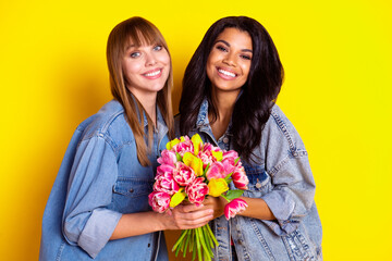 Portrait of two charming cheerful girls arms hold flowers toothy smile isolated on yellow color background