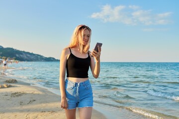 Beautiful teenager with smartphone walking along sea sandy beach at sunset