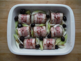 meat rolls in bacon with vegetables, olives and spices in a white plate, on a wooden table, the cooking process.