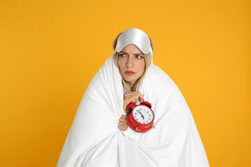 Sad young woman in sleeping mask wrapped with blanket holding alarm clock on yellow background