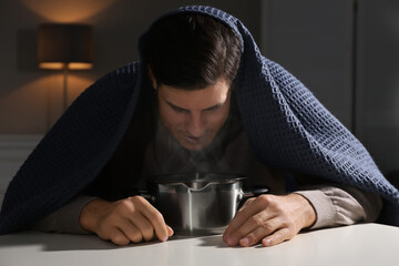 Man with plaid doing inhalation above saucepot at table indoors