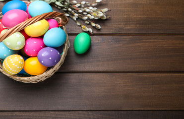 Colorful Easter eggs in wicker basket and willow branches on wooden table, flat lay. Space for text