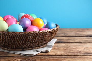 Fototapeta na wymiar Colorful Easter eggs in wicker basket on wooden table against light blue background, closeup. Space for text