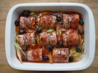 meat rolls in bacon with vegetables, olives and spices in a white plate, on a wooden table, the cooking process.