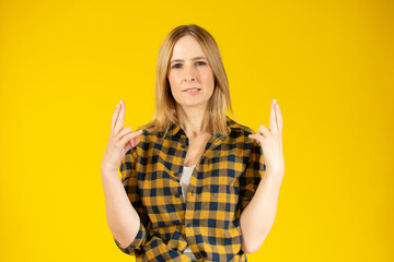 Portrait of a smiling young woman wearing plaid shirt over yellow background, holding fingers crossed for good luck
