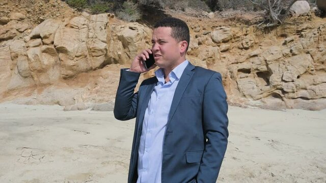 Young businessman in elegant suit talking on the phone while walking on the beach. Remote work in quiet place.