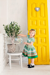 Fototapeta na wymiar Beautiful little blonde girl in floral wreath on white and yellow kitchen background cares for, watering huge plant. Easter concept. Easter card. Color of the year.