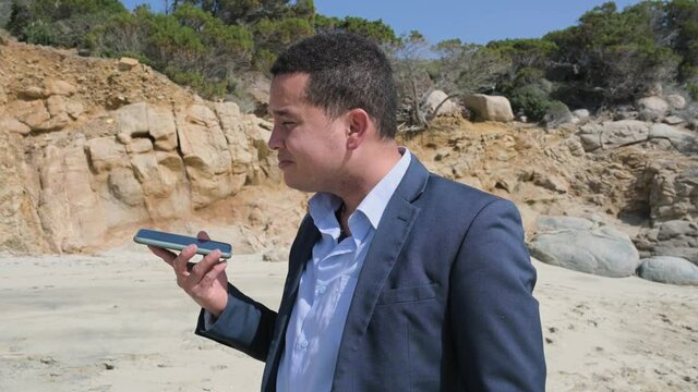 Young businessman in elegant suit using phone listening to a vocal message while walking on the beach. Remote working in a peaceful and quiet place.