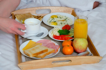 Family on vacations resting in their room. Couple in bathrobes having a breakfast together in bed in hotel, wooden tray closeup. Man and woman are eating and drinking tea. Luxury resting concept.