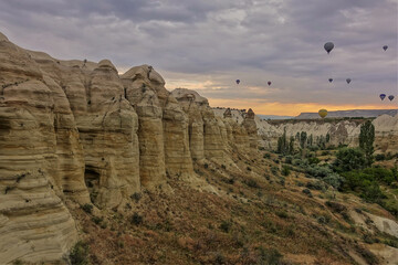 A gorge with vertical slopes similar to stone pillars. Grass and trees grow at the bottom. Colorful balloons soar against the backdrop of a cloudy dawn sky. Cappadocia. Turkey