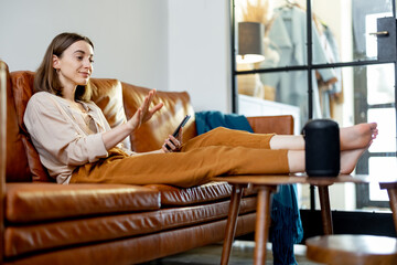 Obraz premium Happy pretty woman speaking with black audio assistant column on the coffee table while sitting on brown leather sofa in stylish living room. Smart home concept.
