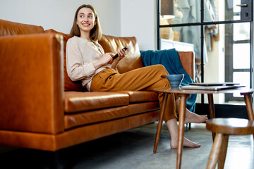 Pretty woman with Smartphone sitting on the brown sofa at home while smiling and looking at the window. Lifestyle and work from home concept. 