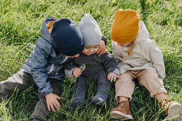 three children, 2 boys and a girl sitting on the green grass dressed in warm spring clothes. Brothers and sister have fun on a walk