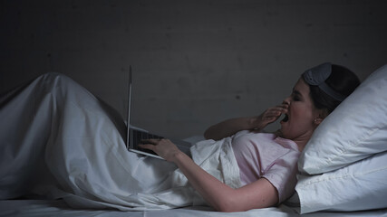 tired freelancer in eye mask using laptop and yawning in bed at night