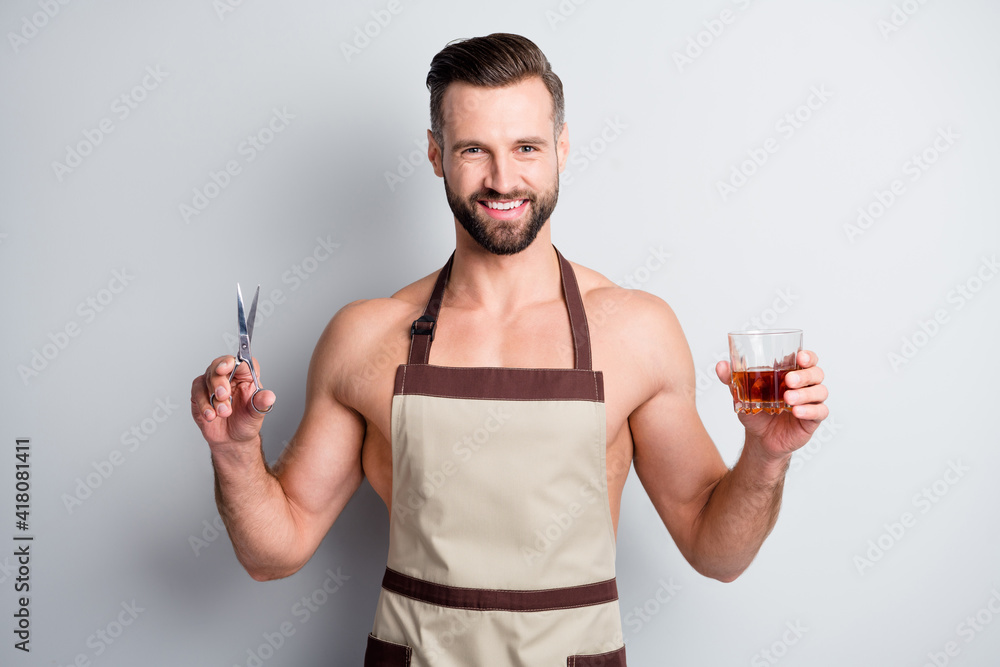 Poster Portrait of attractive cheerful guy wearing apron holding in hands beverage scissors isolated over light grey pastel color background