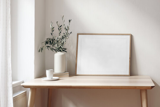 Home office concept. Empty horizontal wooden picture frame mockup. Cup of coffee on wooden table. White wall background. Vase with olive branches. Elegant working space. Scandinavian interior design.