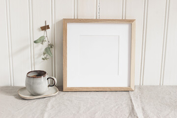 Empty square wooden picture frame mockup. Cup of coffee on linen table cloth. Dry eucalyptus taped...