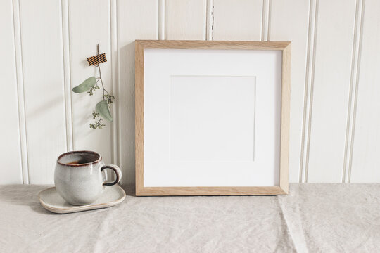 Empty square wooden picture frame mockup. Cup of coffee on linen table cloth. Dry eucalyptus taped on white wooden wall background. Home office concept. Scandinavian interior design.