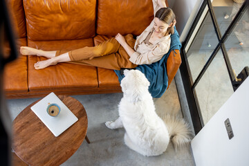 Pretty woman relaxing and dreaming on the brown sofa with a white dog at home. Lifestyle and home coziness concept. Top view.