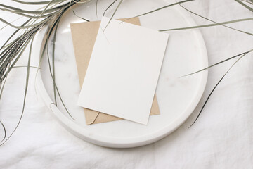 Summer tropical stationery still life scene. Dry date palm leaf on round marble tray. White table cloth in sunlight. Blank greeting card, invitation mockup with craft paper envelope. Vacation flat lay