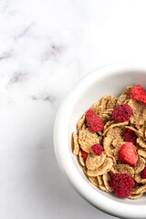 Aerial view of half a bowl with cereals and red berries on a white marble table, vertically, with copy space