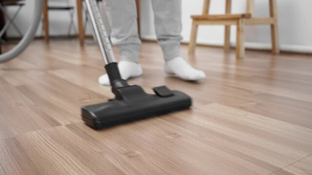 A guy in white socks cleans the floor with laminate with a vacuum cleaner.