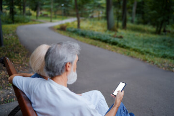 Senior family couple using smartphone together on bench at summer park.
