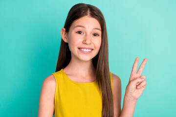 Photo of optimistic nice brown hair girl show v-sign wear yellow dress isolated on bright teal color background