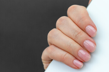Obraz premium Female hand with beautiful manicure - pink nude nails with white smartphone on gray blurred background, closeup with copy space. Selective focus