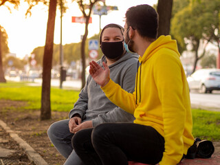 Amigos sentados en un banco en la calle hablando vistiendo mascarilla respetando las medidas de seguridad y distanciamiento social de la pandemia del coronavirus