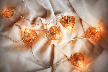 A glowing garland with heart-shaped bulbs glows with a warm light and lies on a white soft fabric,...