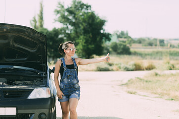 A girl in a denim jumpsuit next to a car with the hood open holds her finger up trying to stop the car
