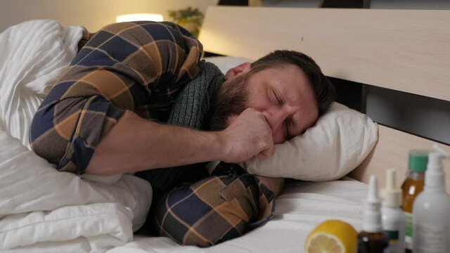 A sick person lies in bed at home, he has a severe dry cough tracheitis, signs of infection. The man covers his mouth with his fist. Season of colds, fever.
