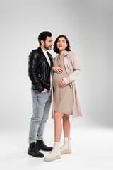 Trendy pregnant woman looking at camera near husband on grey background