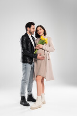 Smiling pregnant woman with flowers standing near stylish husband on grey background