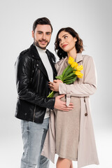 Smiling man in leather jacket hugging pregnant wife with tulips on grey background