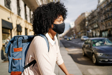 African American woman wearing a protective mask while riding a bicycle in the city. Woman holds backpack on her back