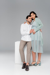 Young man embracing stylish pregnant wife near white cube on grey background