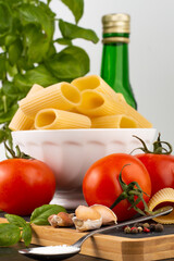 In the foreground is a blurred image of pepper, cloves of garlic, a spoon with salt, a tomato, in the background is a white plate with rigatoni, basil .