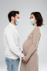 Side view of pregnant woman in medical mask holding hands of husband isolated on grey