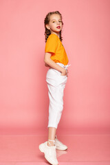 Fototapeta na wymiar full length little girl on pink studio background. wearing an orange T-shirt and white pants and white sneakers. Stylish kids, happy childhood, emotions. copy space