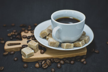 A coffee mug with hot coffee poured into it stands on a white saucer . Coffee beans and lump sugar are scattered nearby .